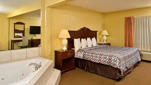 A hotel room with a large bathtub, a bed with a patterned bedspread, two bedside lamps, a nightstand, a dresser, a mirror, and a television.