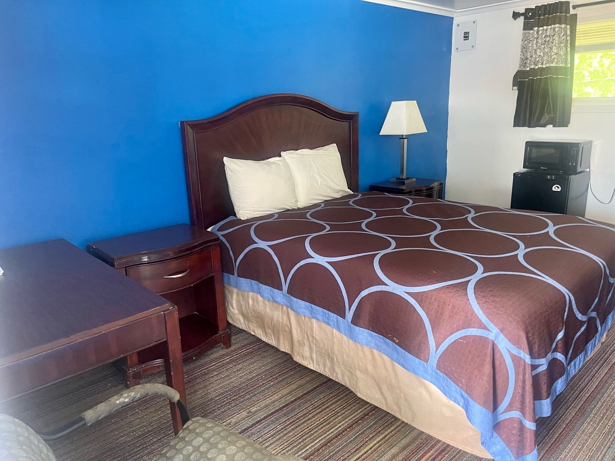 Habitación de hotel con cama, escritorio, silla, mesita de noche, lámpara, microondas y ventana con cortinas.