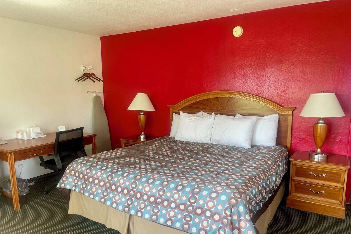 A hotel room with a bed, two bedside tables with lamps, a desk with a chair, and a red accent wall.