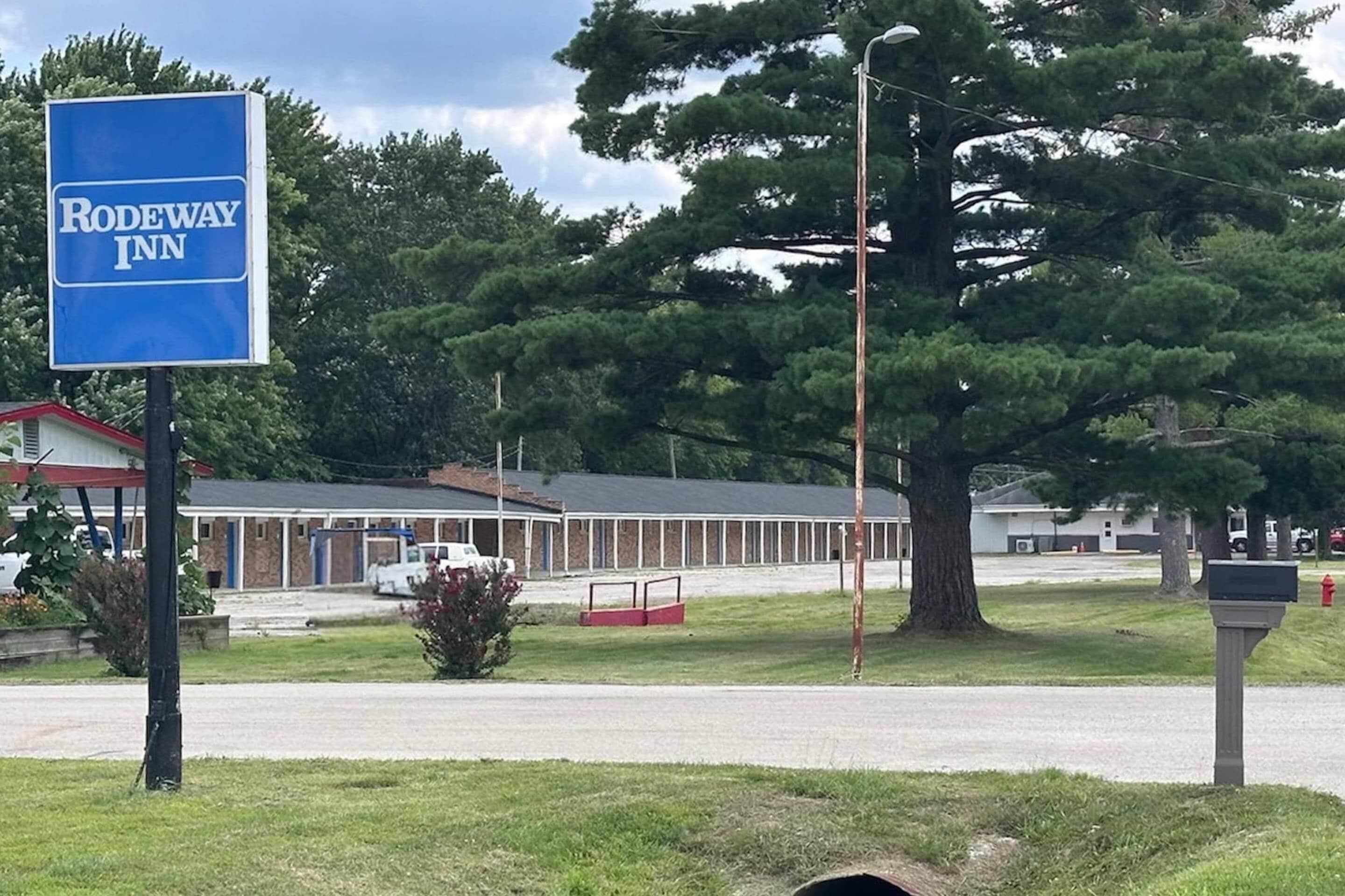 A motel named Rodeway Inn with a large tree in the foreground and a building with a red roof in the background.