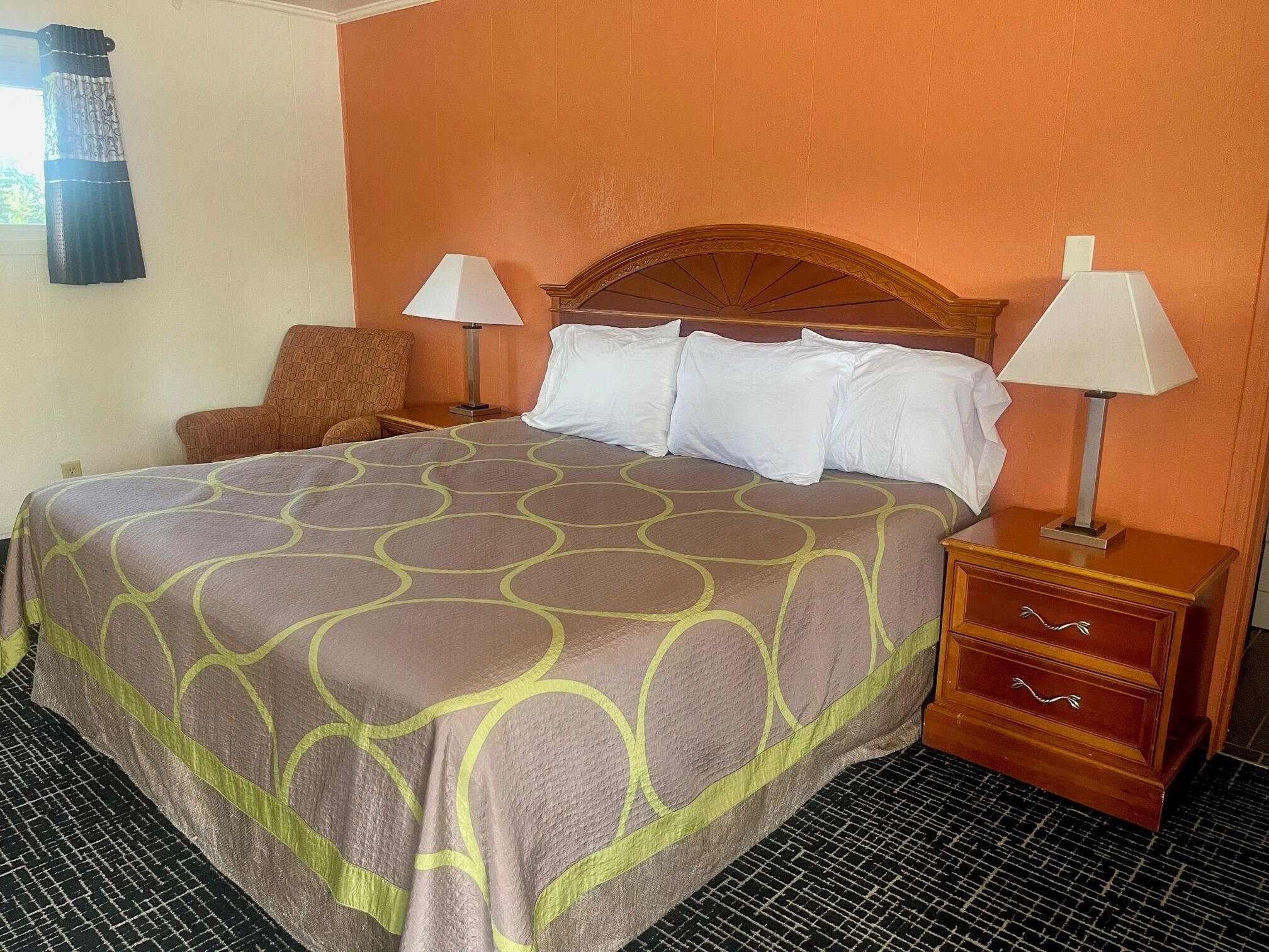 A hotel room with a bed, two bedside lamps, a chair, and a wooden nightstand with drawers.