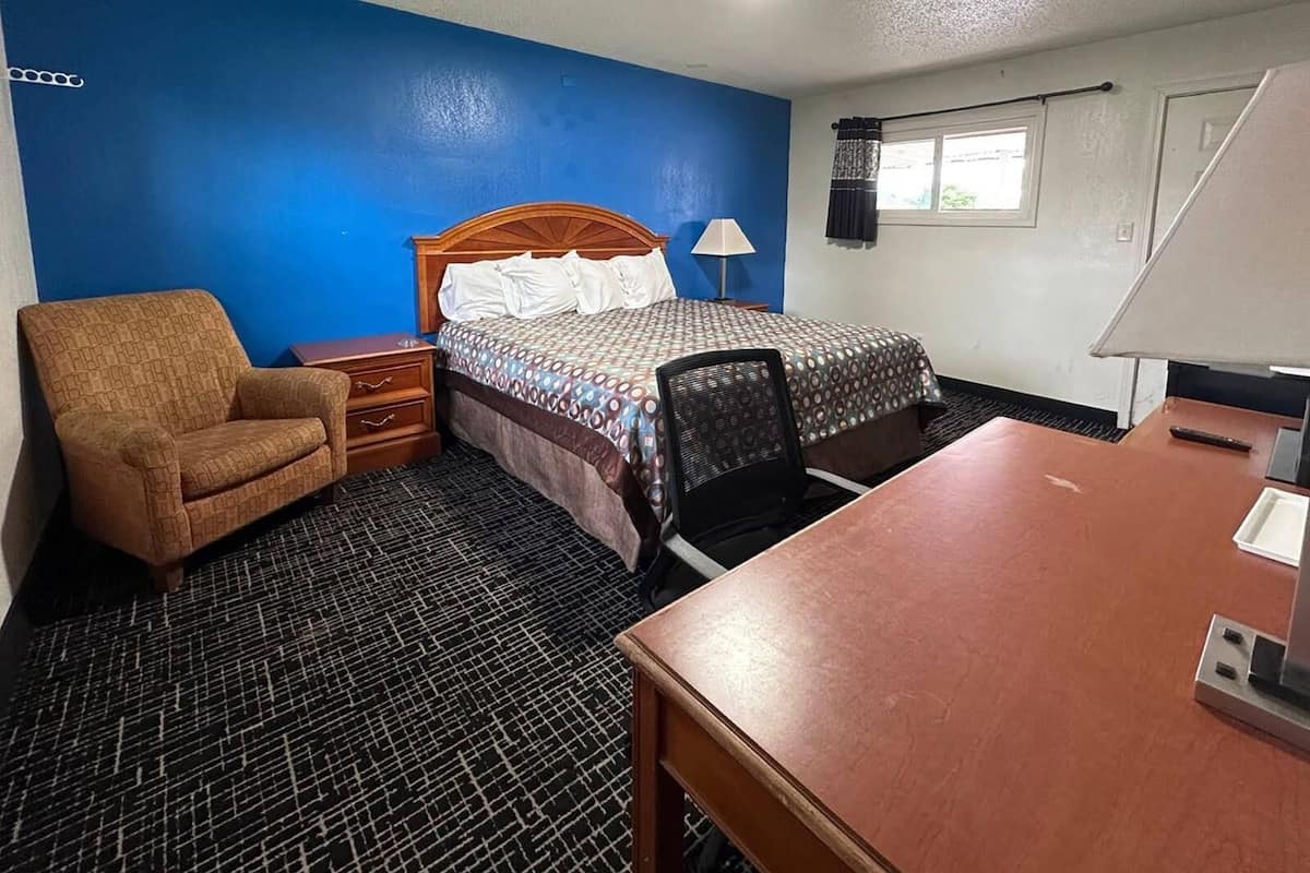 A hotel room with a bed, a desk, a chair, a nightstand, and a lamp.