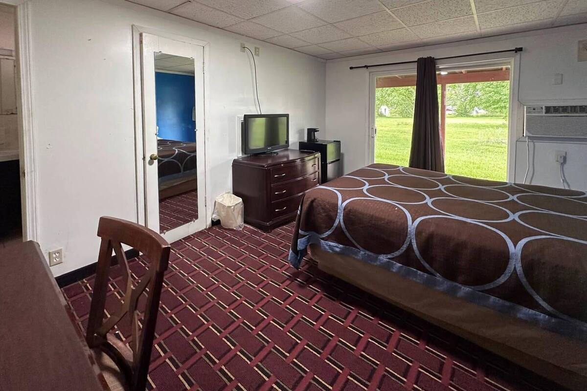 A hotel room with a bed, a desk, a chair, a television, and a window with a view of a grassy area.