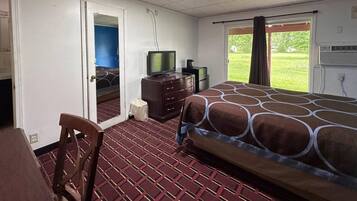 A hotel room with a bed, a desk, a chair, a television, and a window with a view of a grassy area.