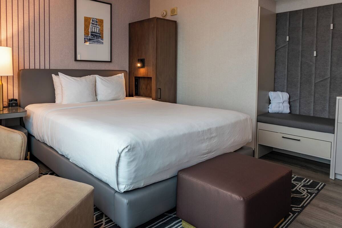 Ein Hotelzimmer mit einem großen Bett, einem Sessel, einer Bank, einem Nachttisch mit Lampe, einem Schrank und einem gerahmten Bild an der Wand.