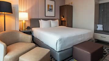 Habitación de hotel con una cama grande, una silla, un banco, una mesita de noche con lámpara, un armario y un cuadro en la pared.