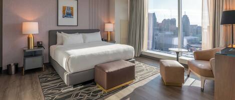 A modern hotel room with a large bed, two bedside tables with lamps, a small table, a sofa, and a view of the city skyline.
