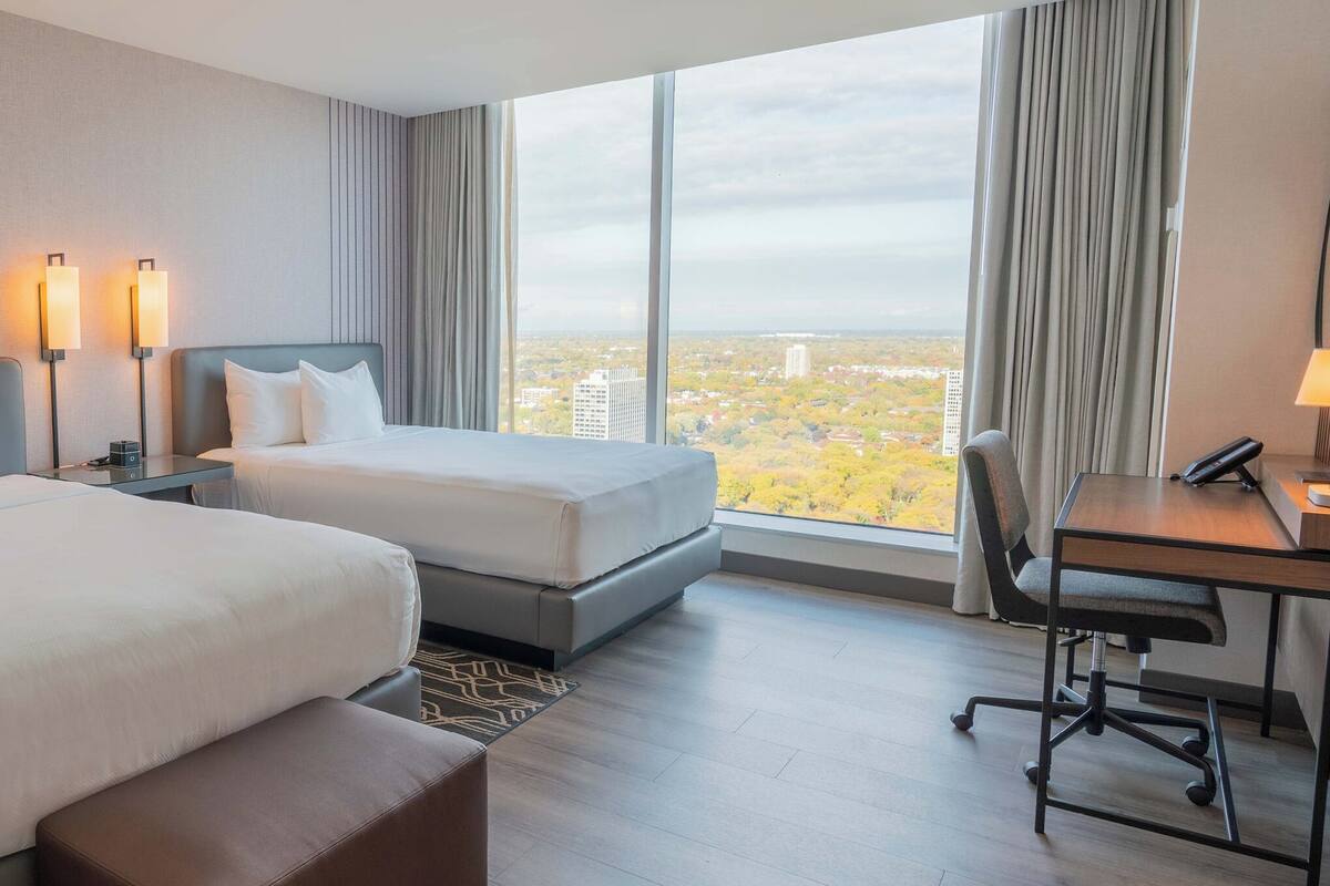 Ein Hotelzimmer mit zwei Betten, einem Schreibtisch und einem Sessel. Ein großes Fenster bietet Blick auf die Stadt.