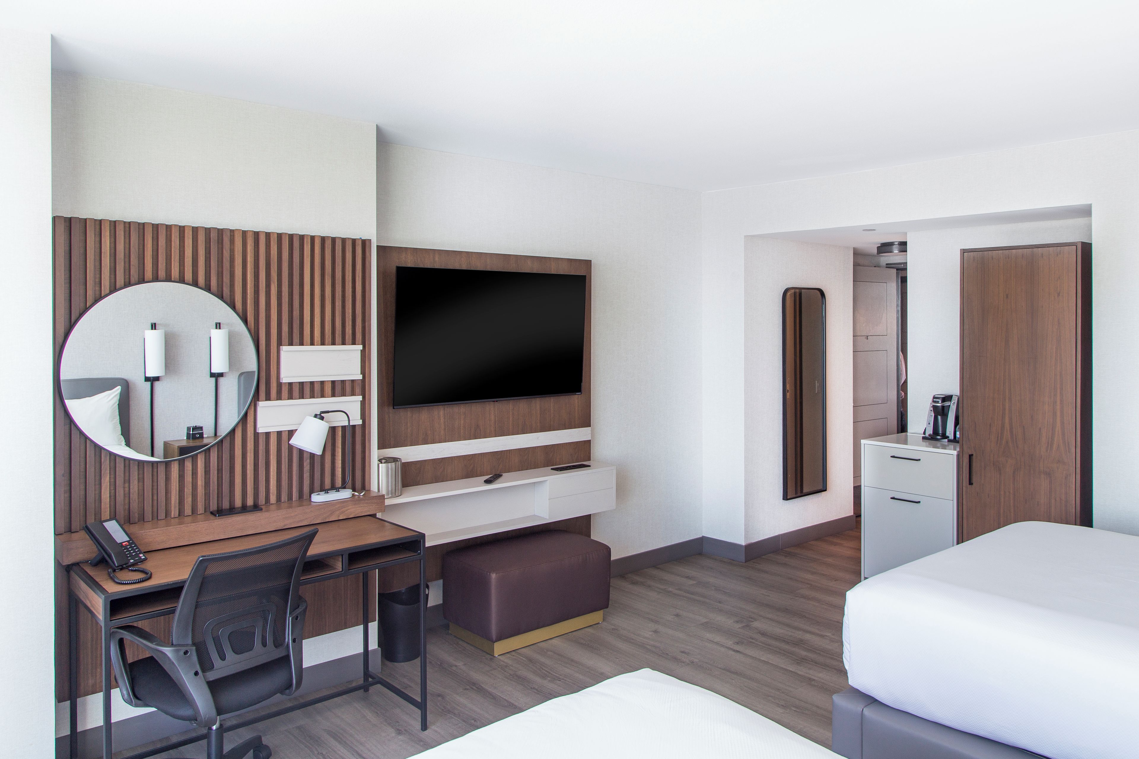 A hotel room with a bed, a desk with a chair, a television, a mirror, and a wardrobe.