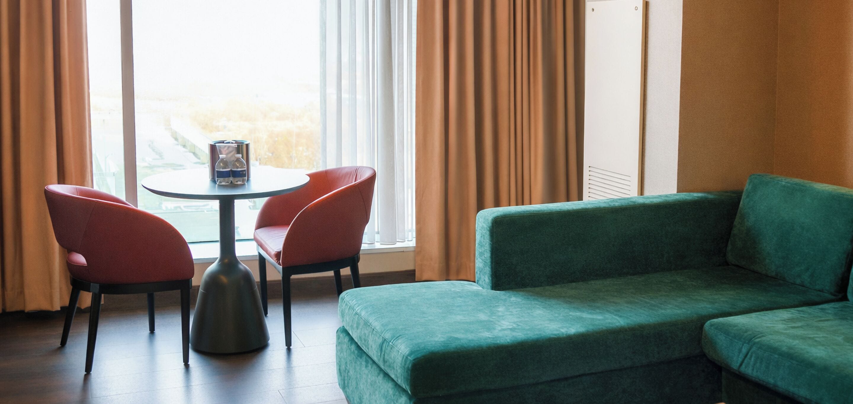 A room with a green sofa, two red chairs, a small round table with a water bottle, and a window with curtains.