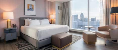 A modern hotel room with a large bed, a seating area with a chair and ottoman, a small table, and a city view through large windows.