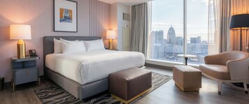 Una habitación de hotel moderna con una cama grande, un área de descanso con un sillón y reposapiés, una mesita y vistas a la ciudad a través de amplios ventanales.