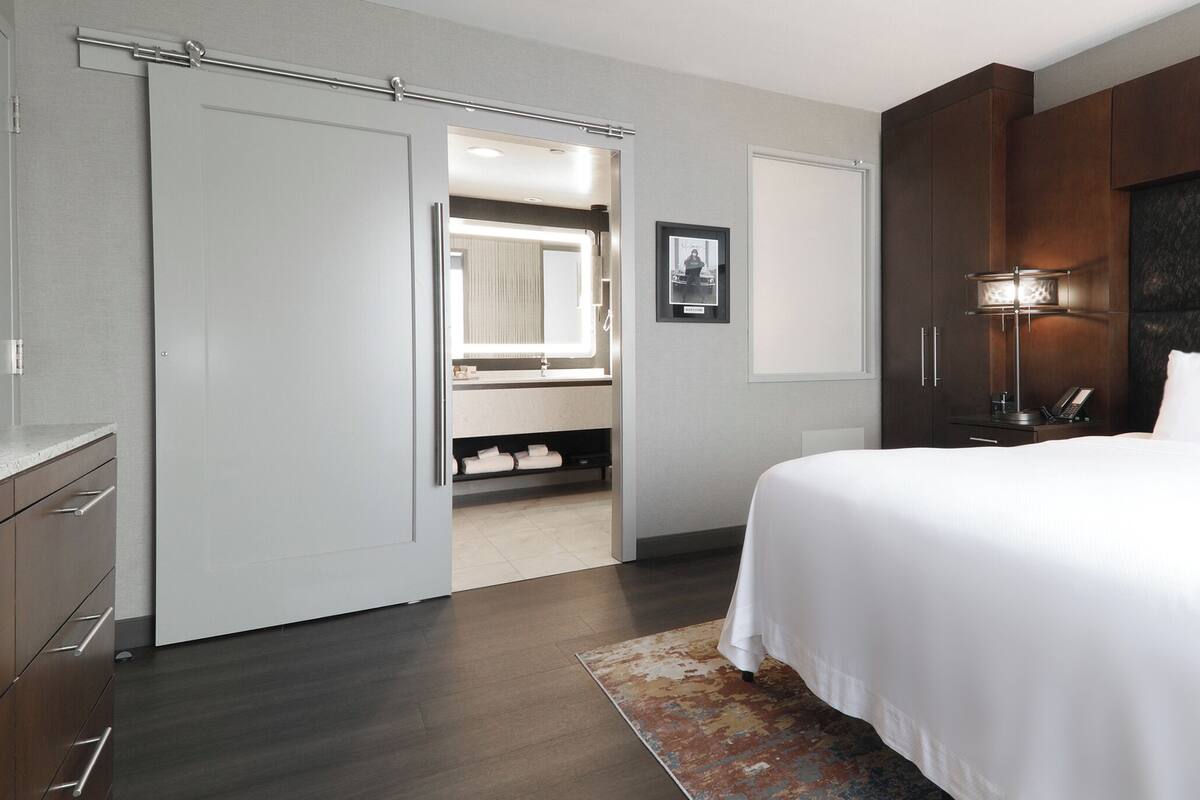Ein modernes Hotelzimmer mit einem großen Bett, einem begehbaren Kleiderschrank, einem Badezimmer mit Spiegel und einem Nachttisch mit Lampe.
