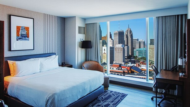 A hotel room with a large bed, a desk, a chair, and a view of the cityscape. - Hollywood Casino at Greektown (Detroit)