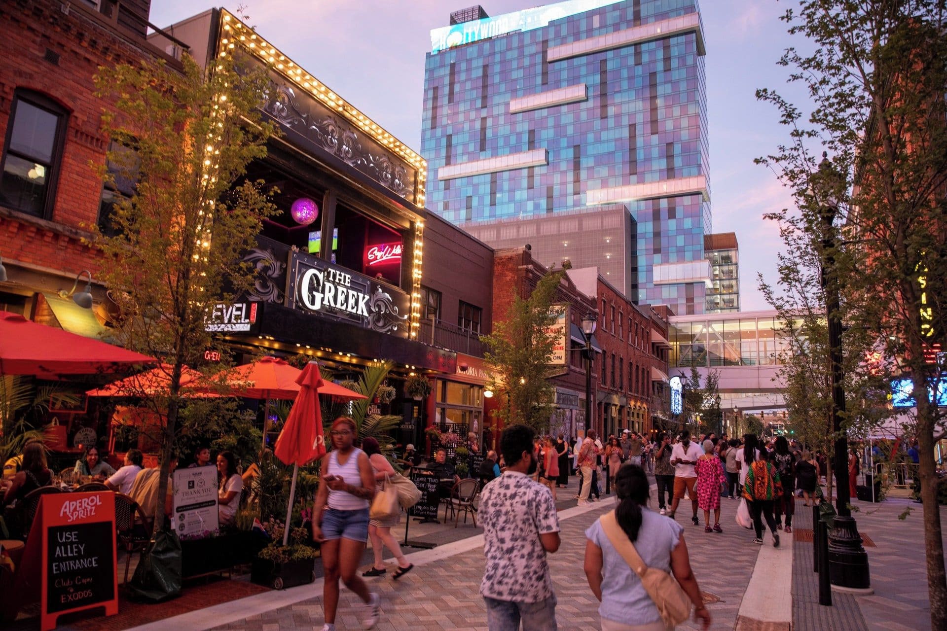 A busy urban street with a variety of shops and restaurants, including a Greek restaurant.