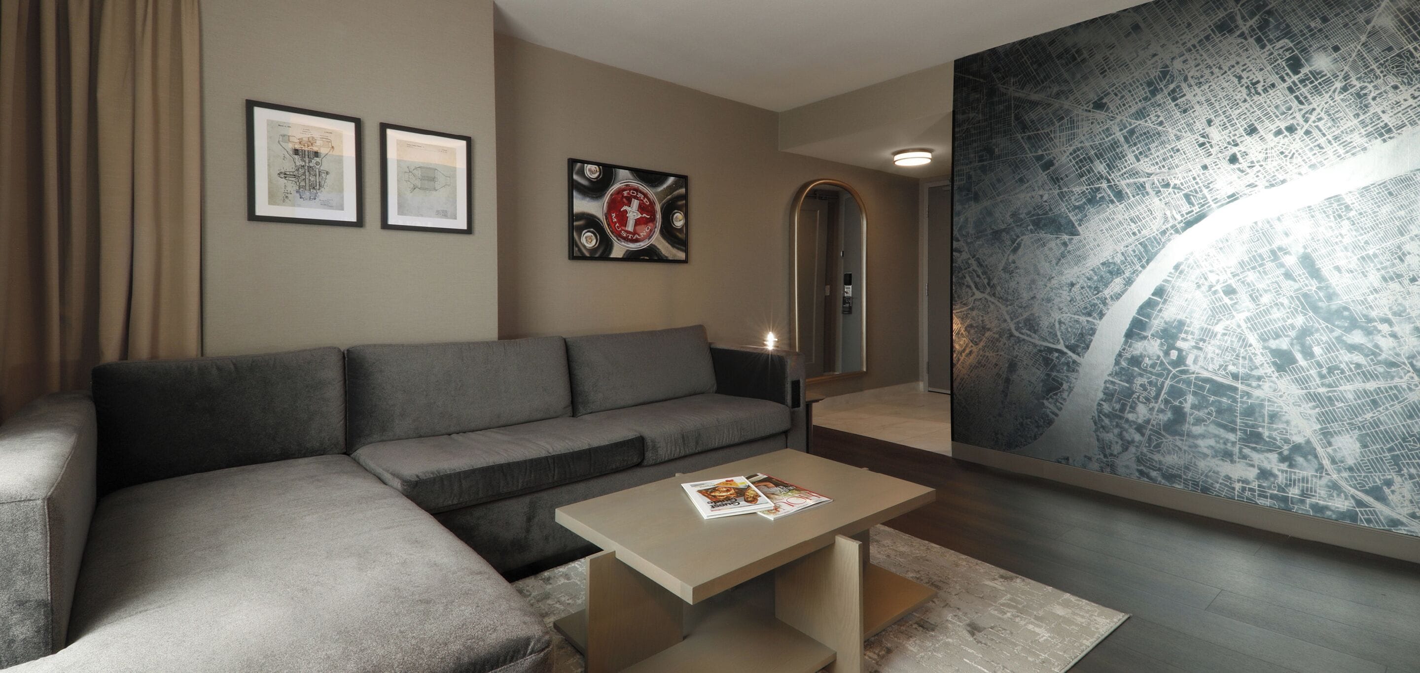 A modern living room with a grey sectional sofa, a coffee table with a magazine, framed artwork on the wall, and a large abstract cityscape mural.