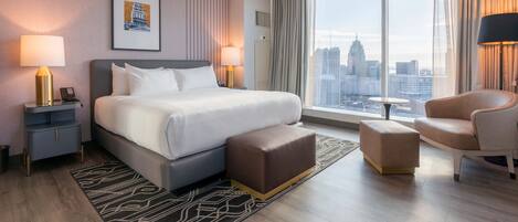 Una habitación de hotel moderna con una cama grande, una zona de estar con una silla y un reposapiés, una mesita pequeña y un ventanal con vistas a la ciudad.