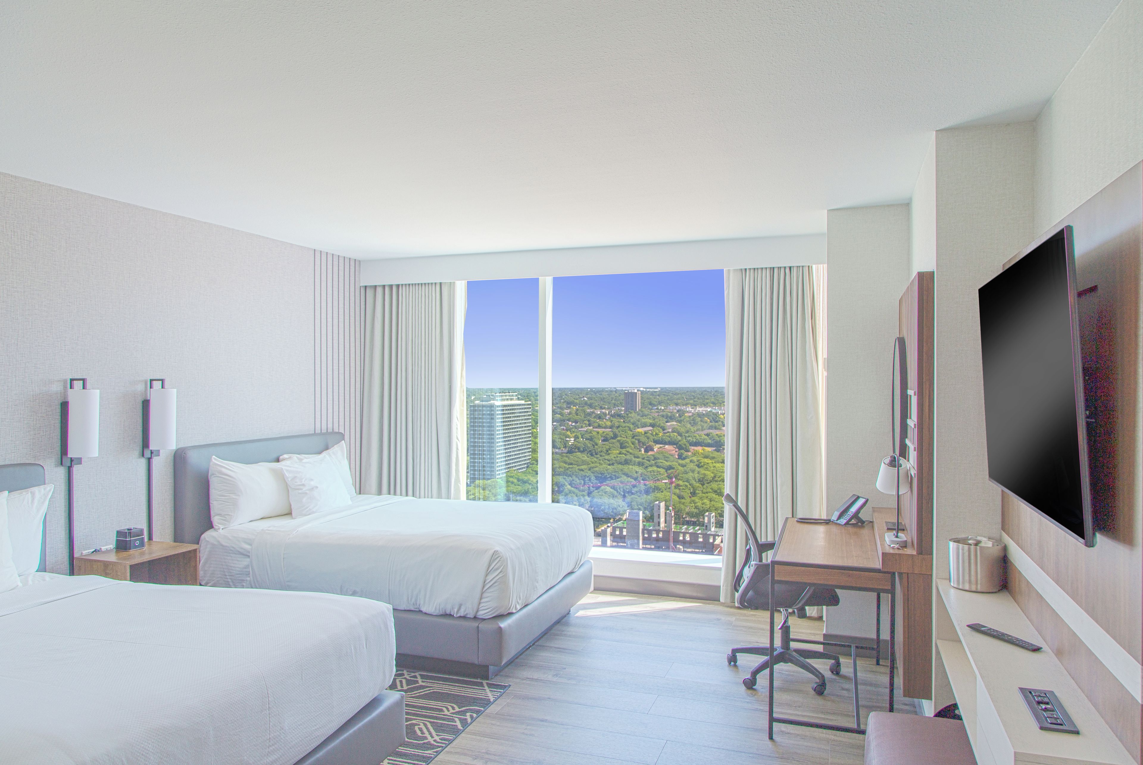 A hotel room with two beds, a desk, a TV, and a view of a cityscape.