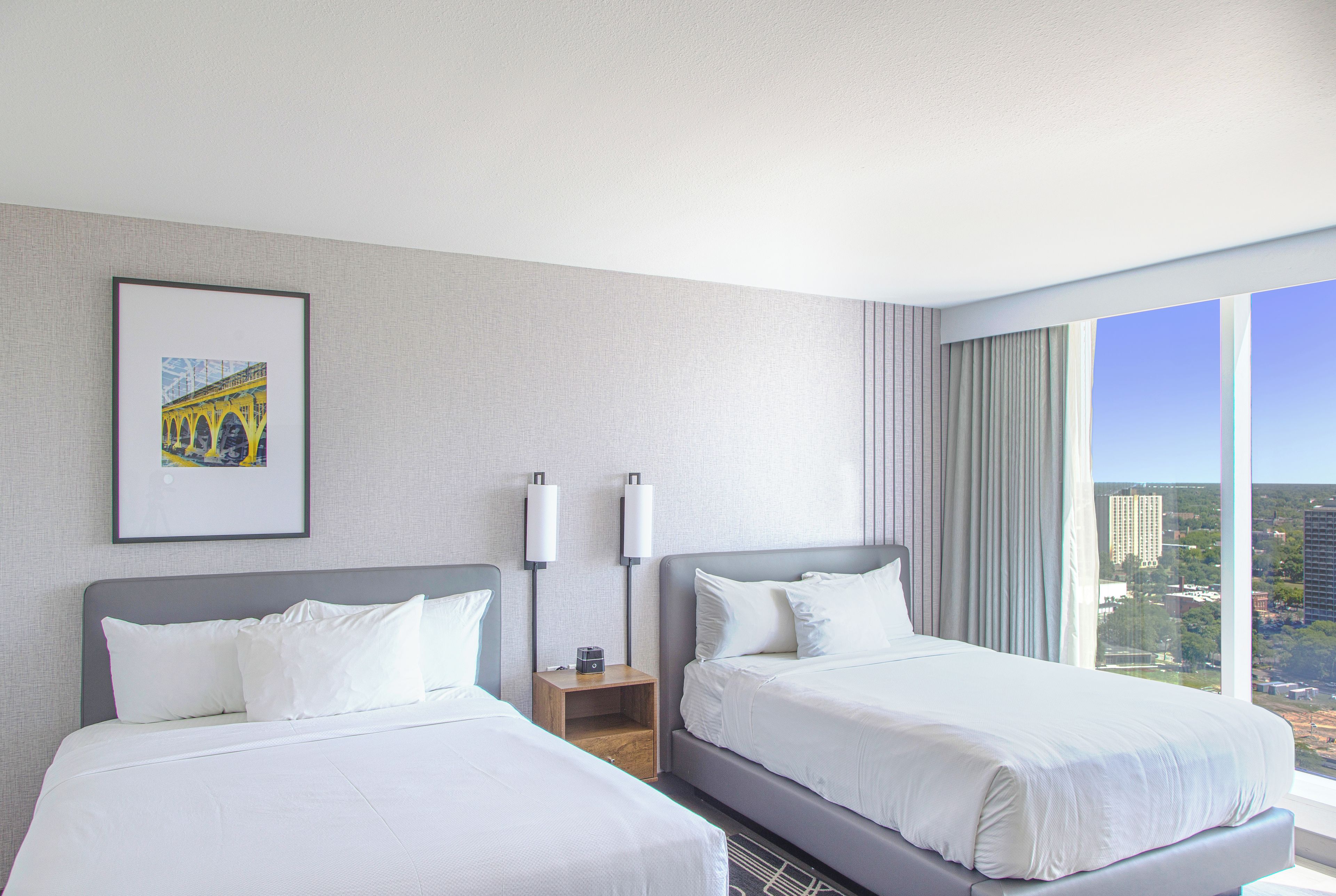 A hotel room with two beds, a nightstand, a framed picture on the wall, and a view of the city through the window.
