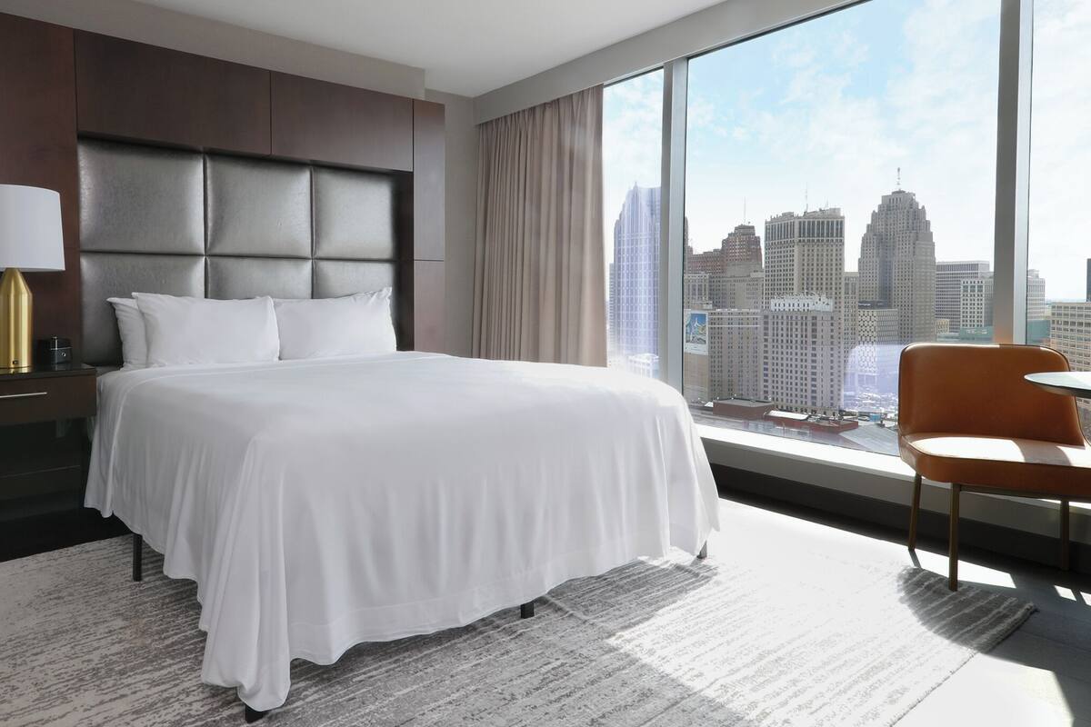 Ein modernes Hotelzimmer mit einem großen Bett, einem Nachttisch, einer Lampe, einem Sessel und Blick auf die Stadtlandschaft.