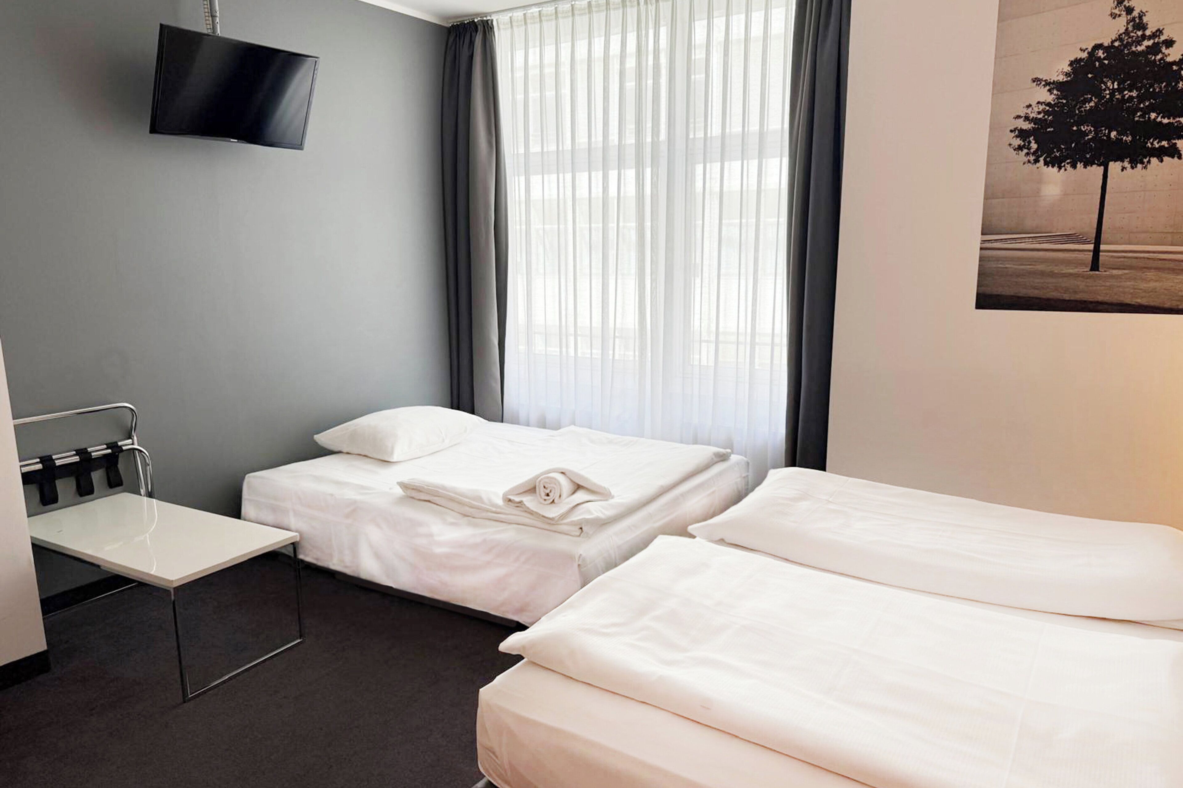 A hotel room with two beds, a flat-screen TV mounted on the wall, a small table, and a window with sheer curtains.