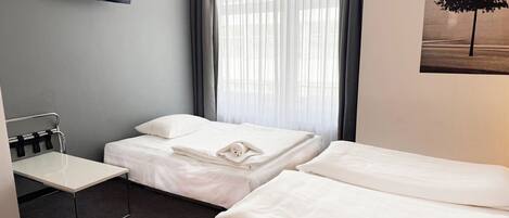 A hotel room with two beds, a flat-screen TV mounted on the wall, a small table, and a window with sheer curtains.