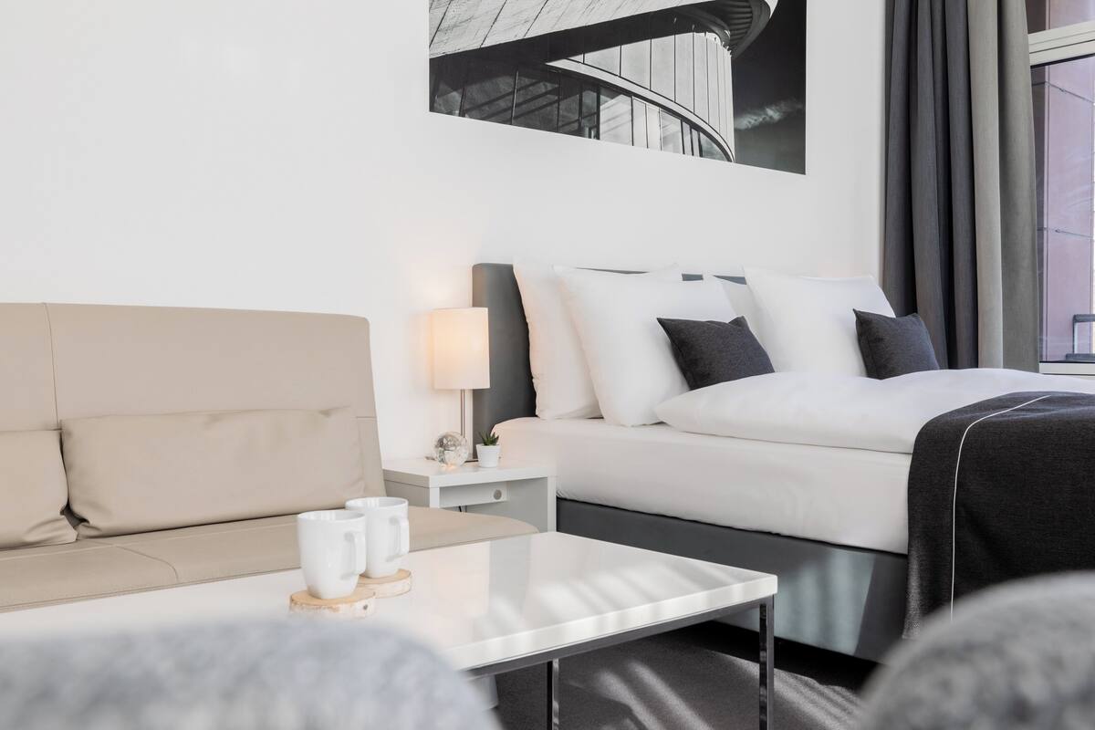 Una habitación de hotel moderna con cama, sofá, mesita de noche con lámpara y una mesa de centro.