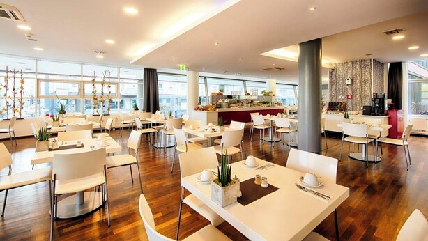 A spacious dining area with multiple tables and chairs, a central bar, and large windows offering views of the city. - Garner Hotel Berlin - Gendarmenmarkt by IHG (Berlin)