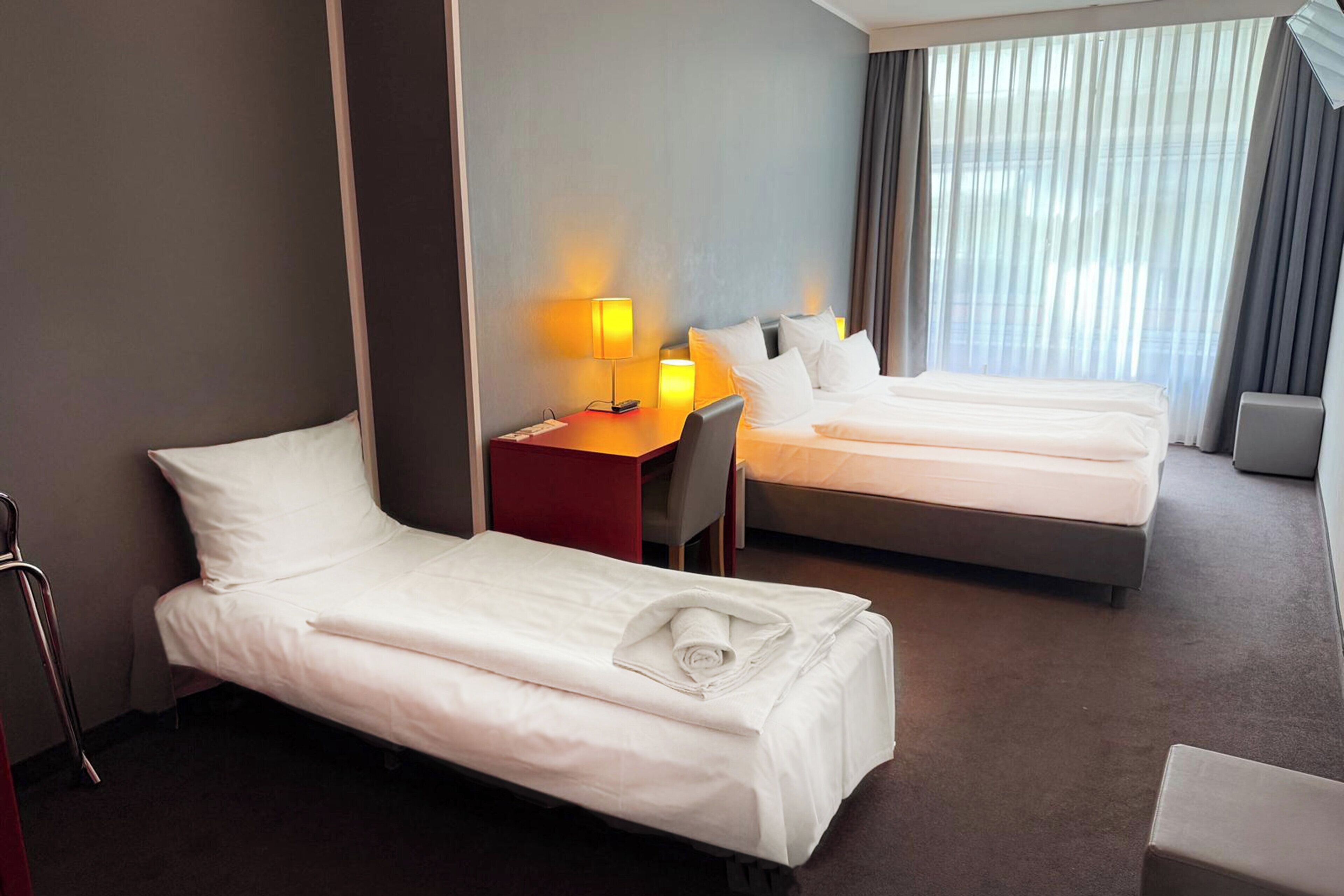 A hotel room with two single beds, a desk, a chair, and a lamp.