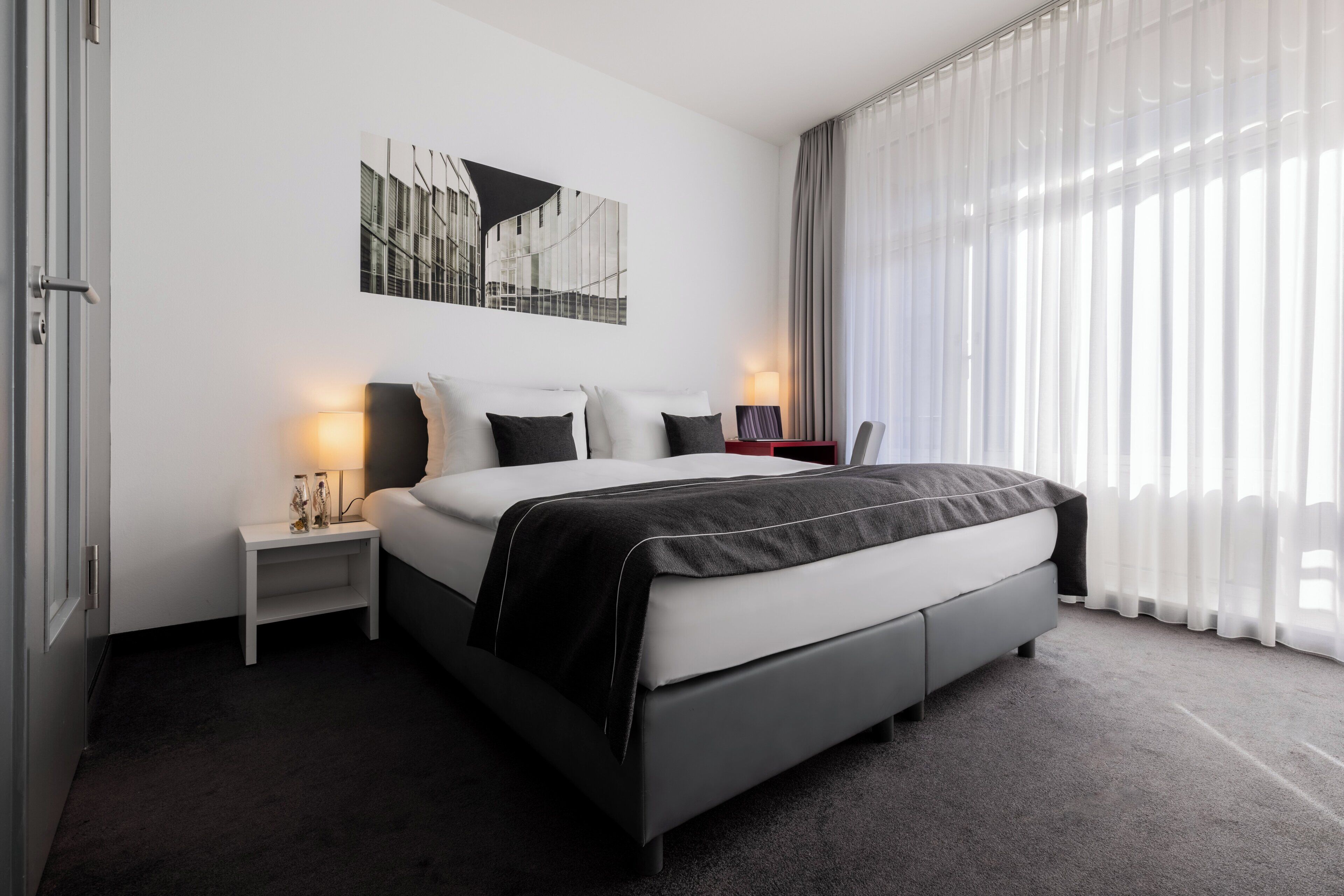 A modern hotel room with a large bed, a nightstand with a lamp, a wall-mounted picture, and sheer curtains allowing natural light.
