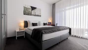 Una habitación de hotel moderna con una cama grande, una mesita de noche con lámpara, un cuadro en la pared y cortinas traslúcidas que dejan entrar luz natural.