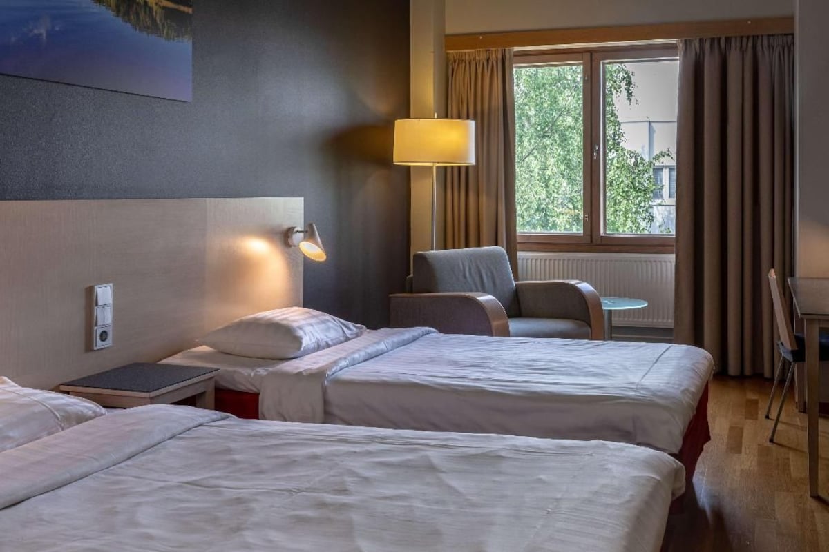 Une chambre d’hôtel avec deux lits, un bureau, une chaise, une lampe et une fenêtre avec des rideaux.