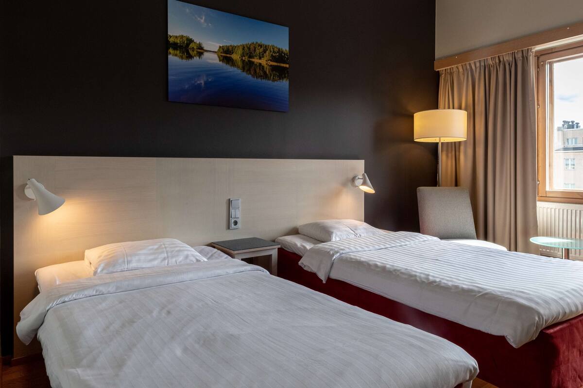 Une chambre d’hôtel avec deux lits, une table de chevet, une lampe, une chaise, une petite table et un tableau au mur.