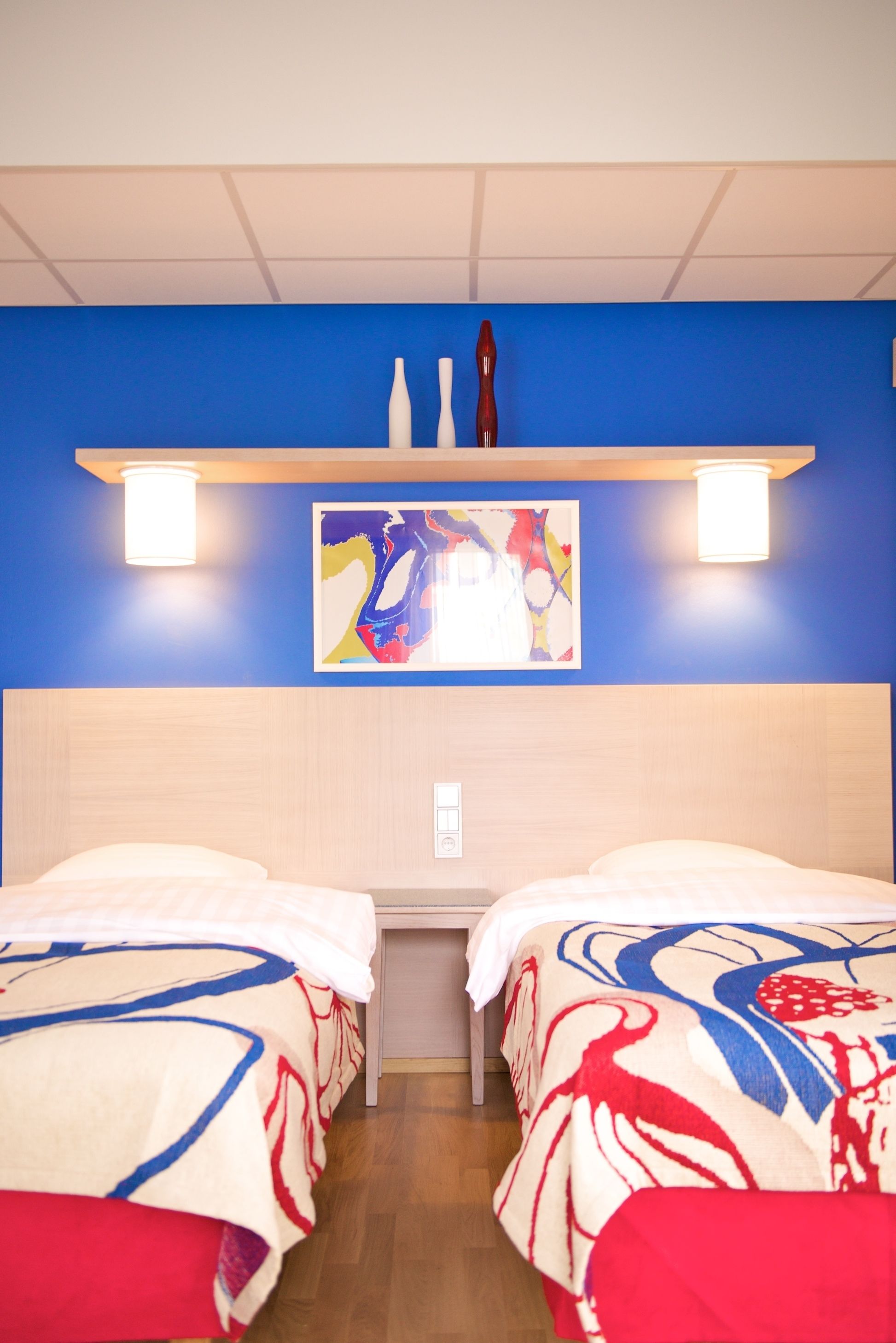 A hotel room with two beds, a blue accent wall, a painting, and a shelf with decorative items.