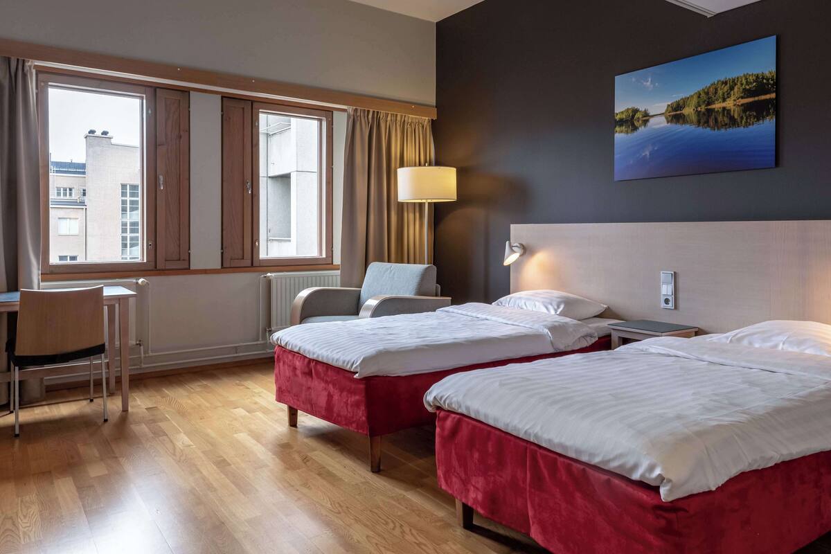 Une chambre d’hôtel avec deux lits, un bureau, une chaise, une lampe et une grande fenêtre avec des rideaux.