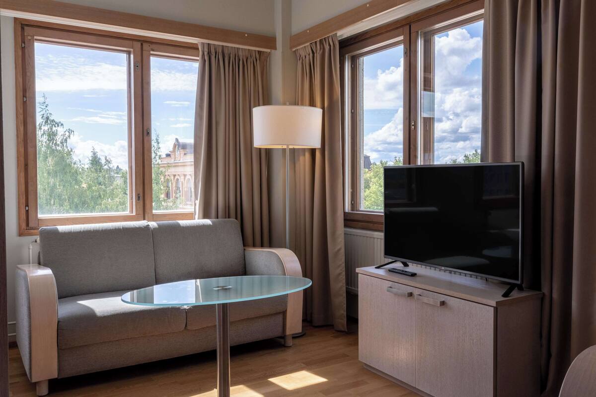 Un salon moderne avec un canapé, une table en verre, un téléviseur posé sur un meuble et de grandes fenêtres avec des rideaux.