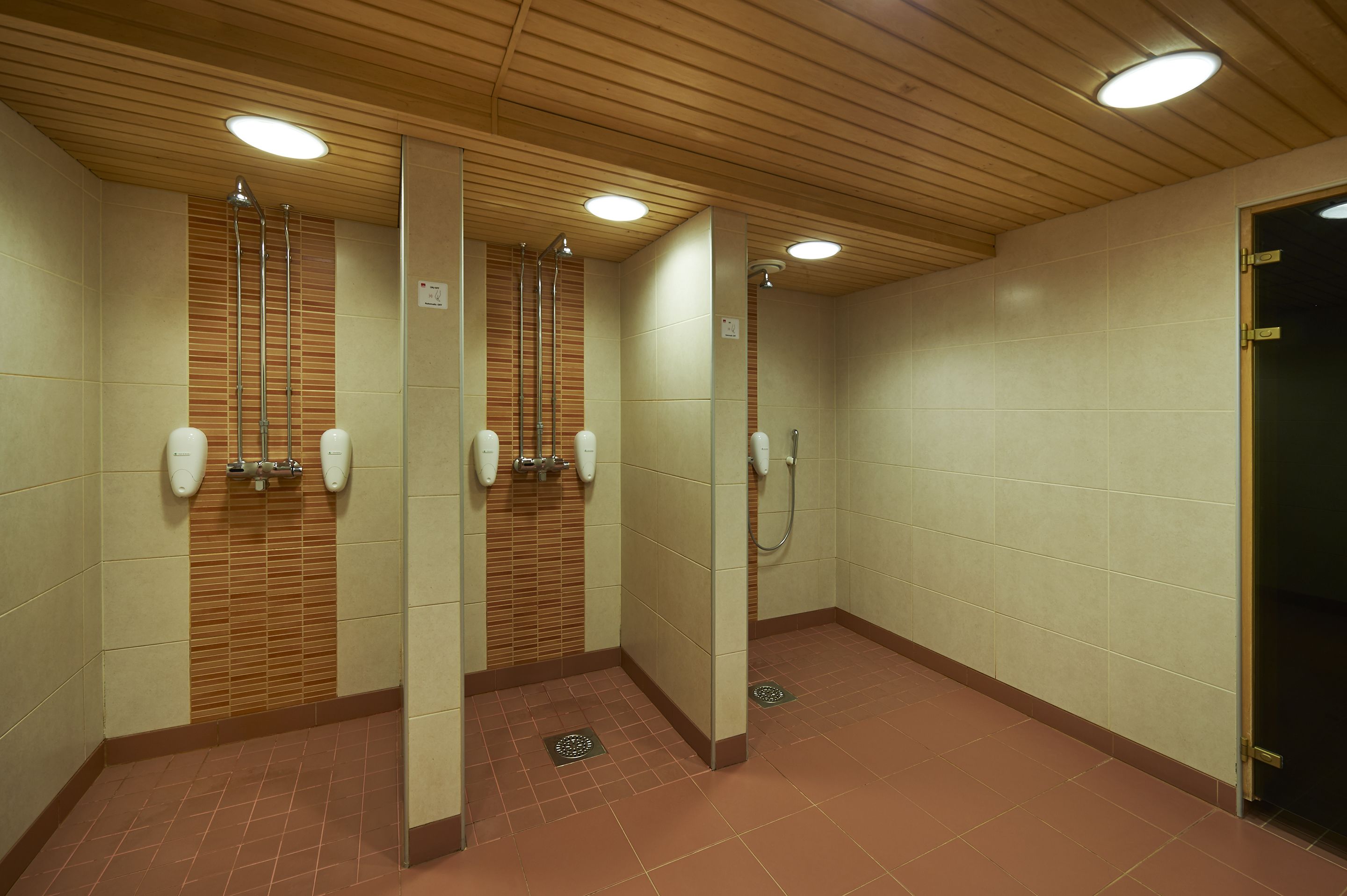 A public restroom with three sinks, each with a soap dispenser and a mirror. The walls are tiled, and the floor is covered with brown tiles.