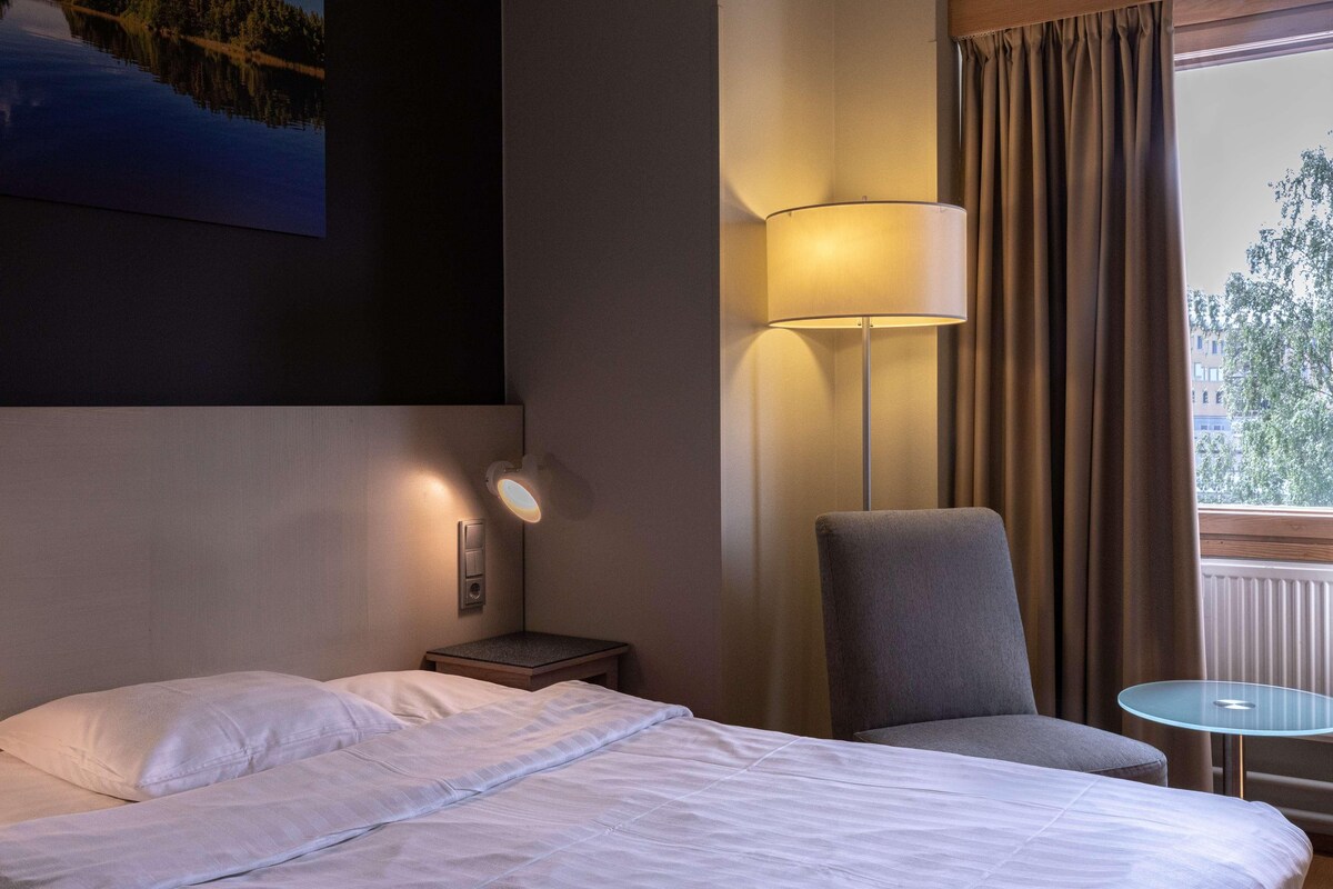 A hotel room with a bed, a chair, a small table, a lamp, and a painting on the wall.