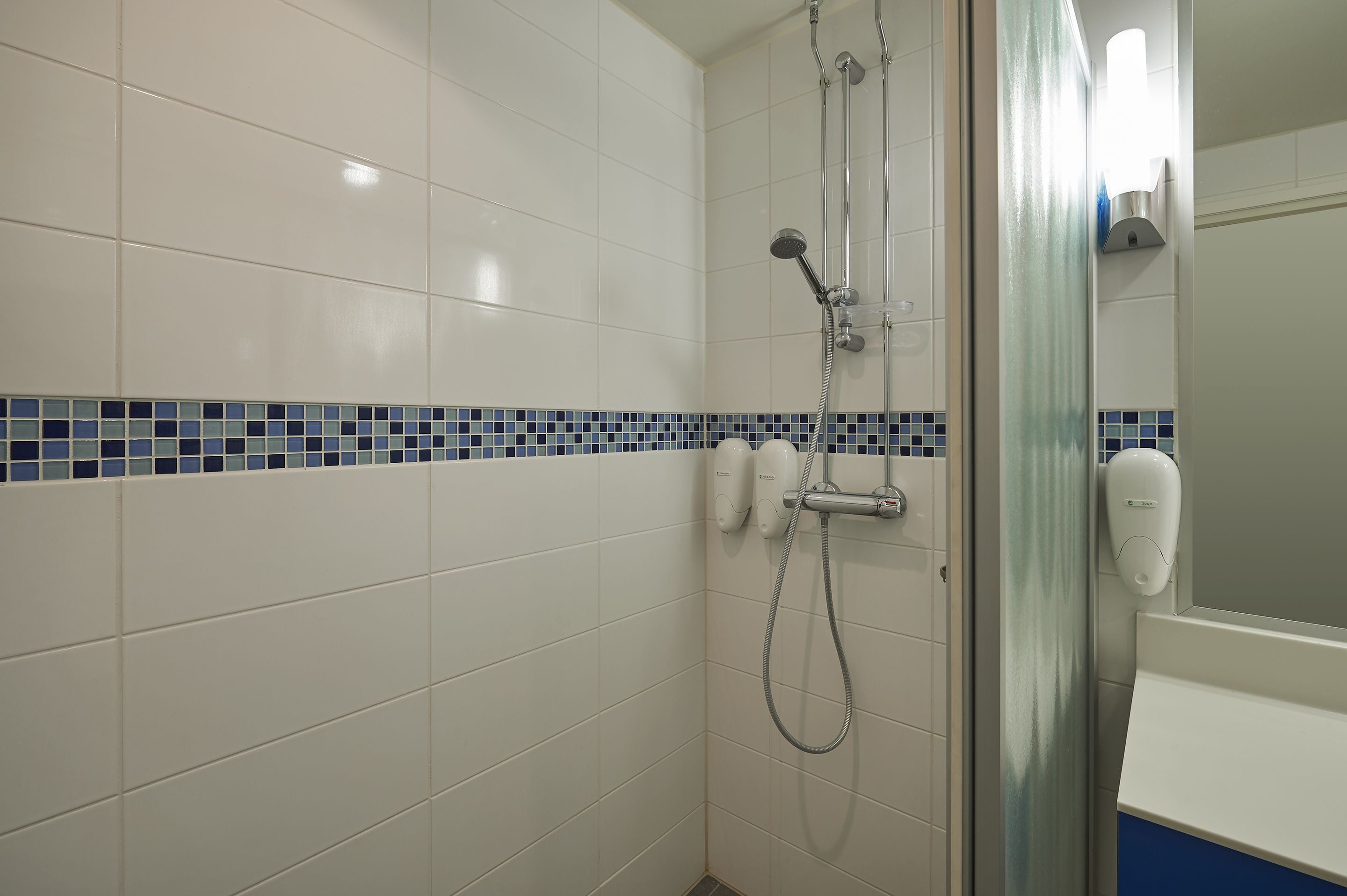 A bathroom with white tiles, a blue-tiled border, a glass shower enclosure, and a hand dryer.