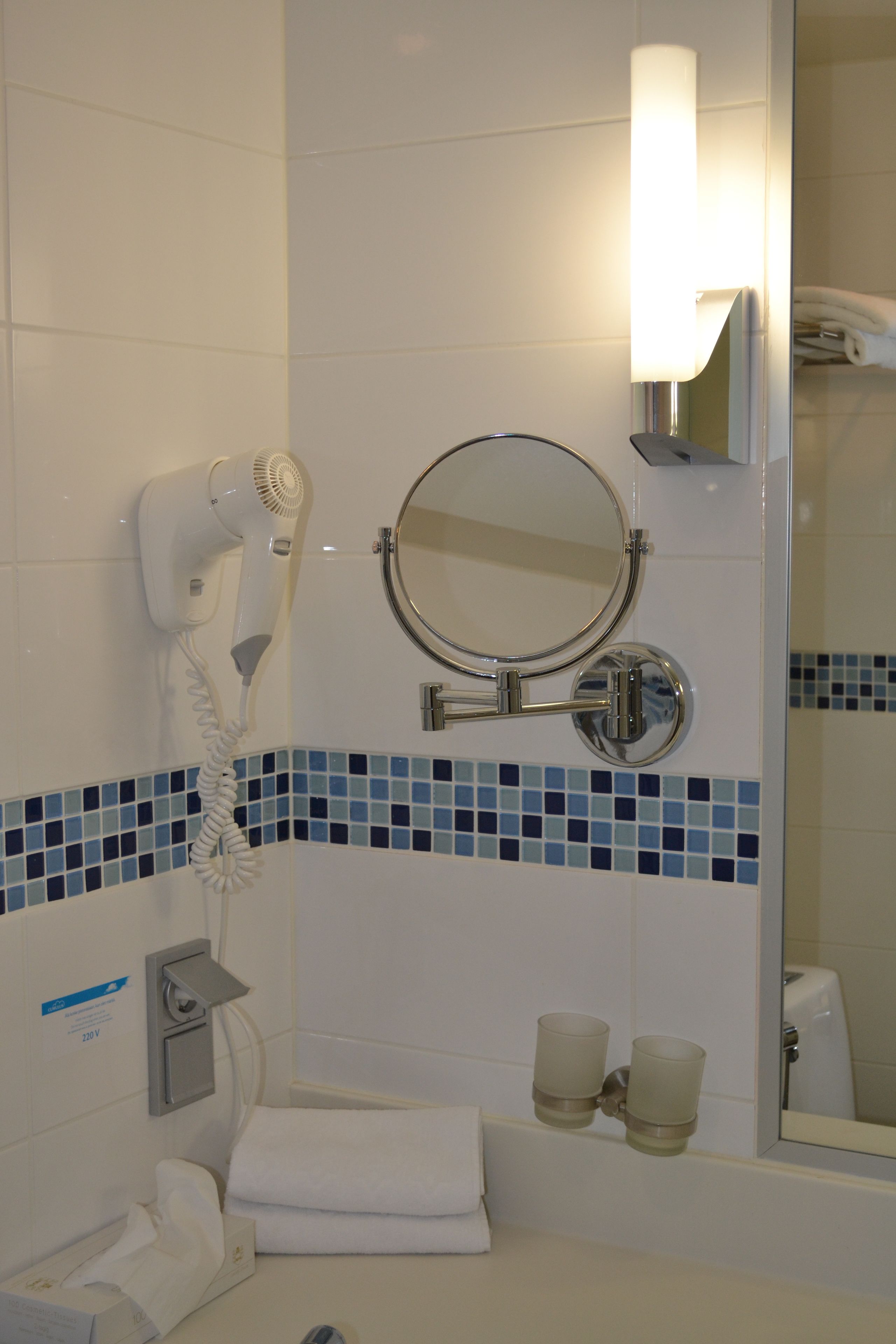 Casa de banho com secador de cabelo, espelho redondo e duas chávenas no balcão.
