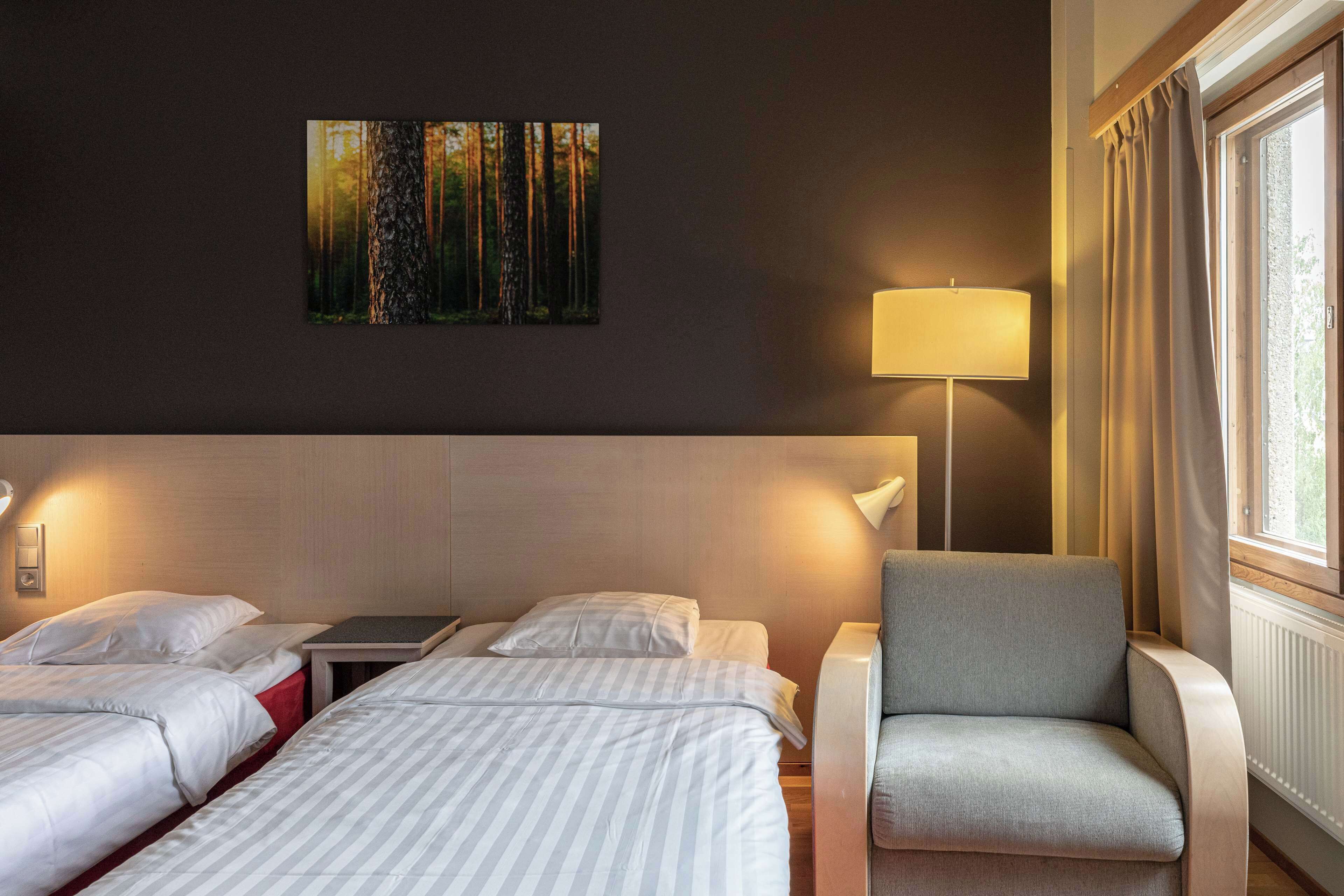 A hotel room with two beds, a chair, a lamp, and a painting of a forest.