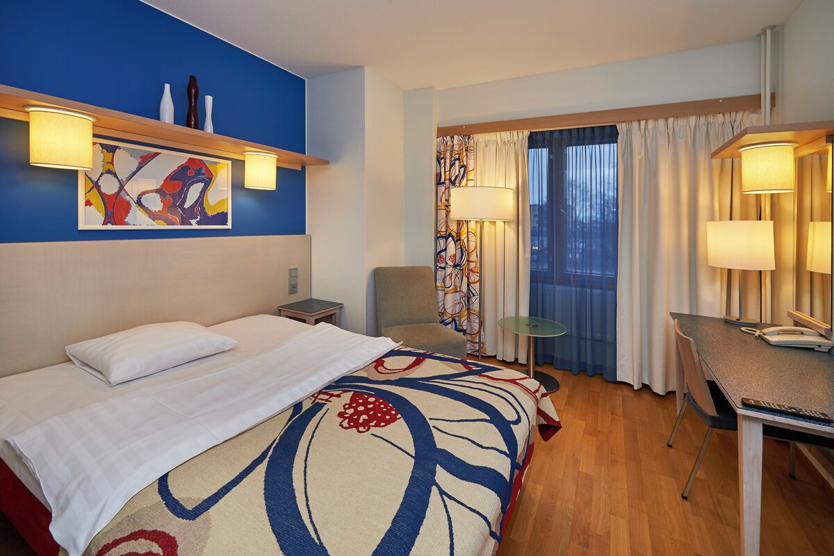 A hotel room with a bed, a desk, a chair, a window with curtains, and a painting on the wall.