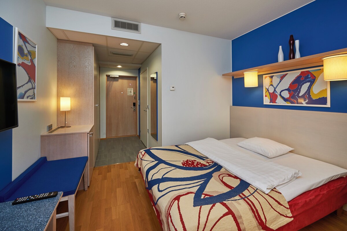 A hotel room with a bed, a desk with a chair, a television, and a painting on the wall.