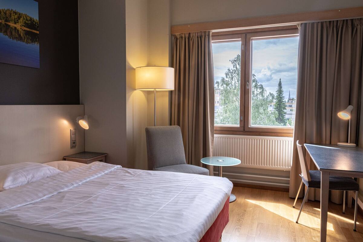Une chambre d’hôtel avec un lit, une chaise, une table, une lampe, une fenêtre avec des rideaux et une vue sur l’extérieur.