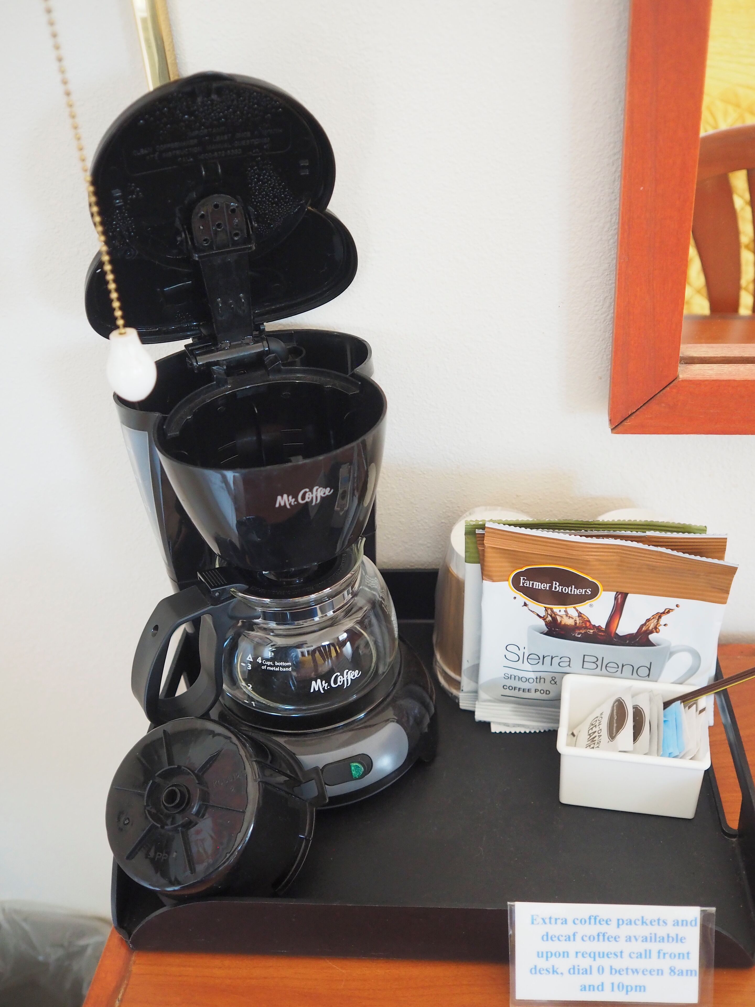 Una cafetera de la marca Mr. Coffee, una bolsa de cápsulas de café Farmer Brothers, y un cartel que indica que se pueden solicitar más sobres de café.