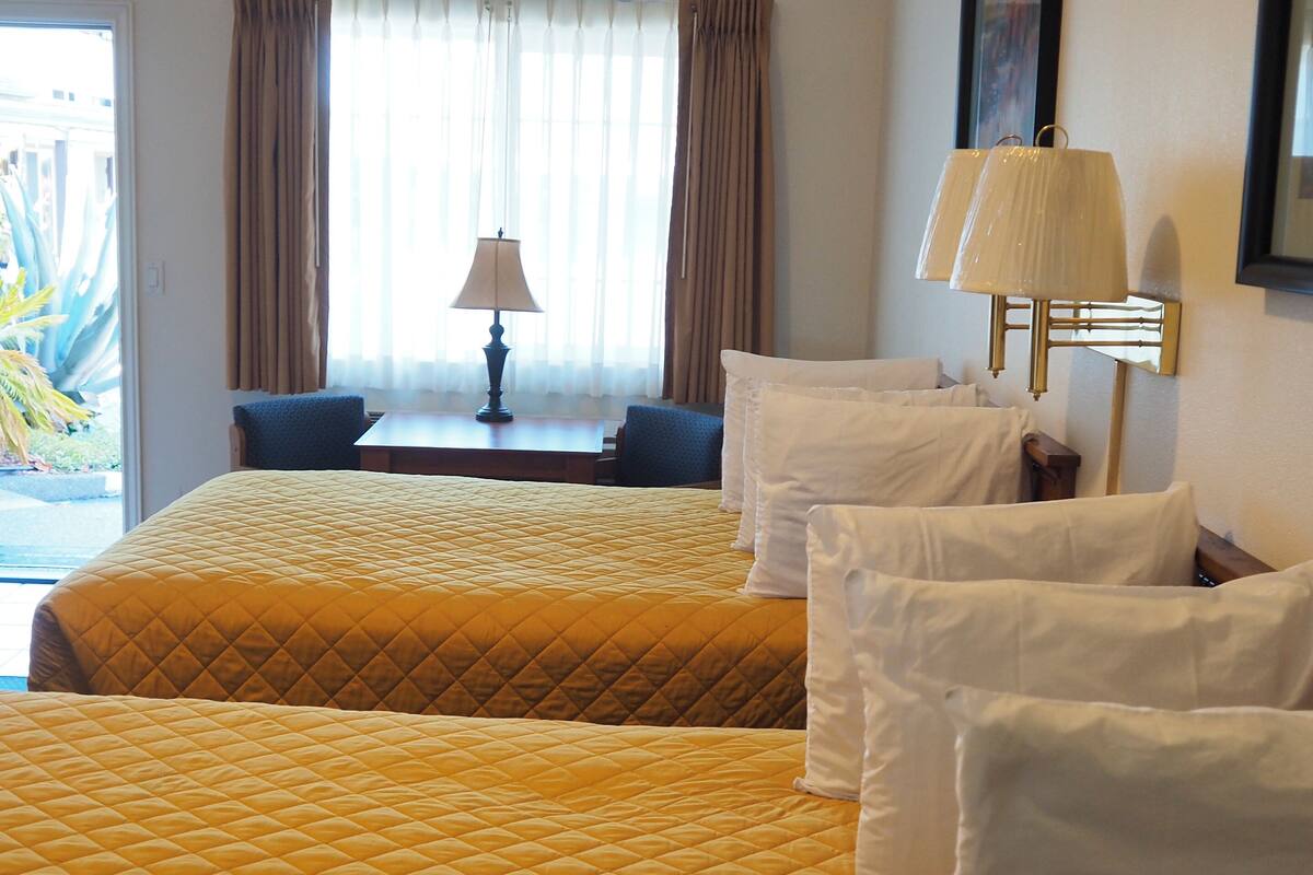 A hotel room with two beds, a desk, a lamp, and a view of the outdoors.