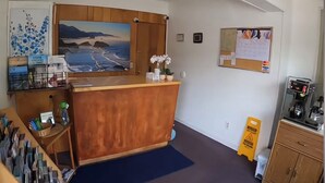 A reception area with a wooden desk, a painting of a coastal landscape, a coffee machine, and a caution sign on the floor.