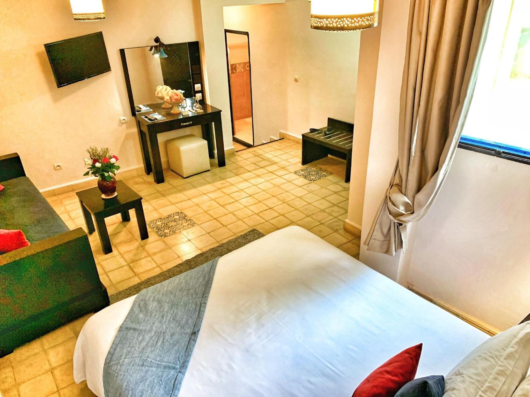 A hotel room with a large bed, a desk with a chair, a small table with a vase, and a TV on the wall.