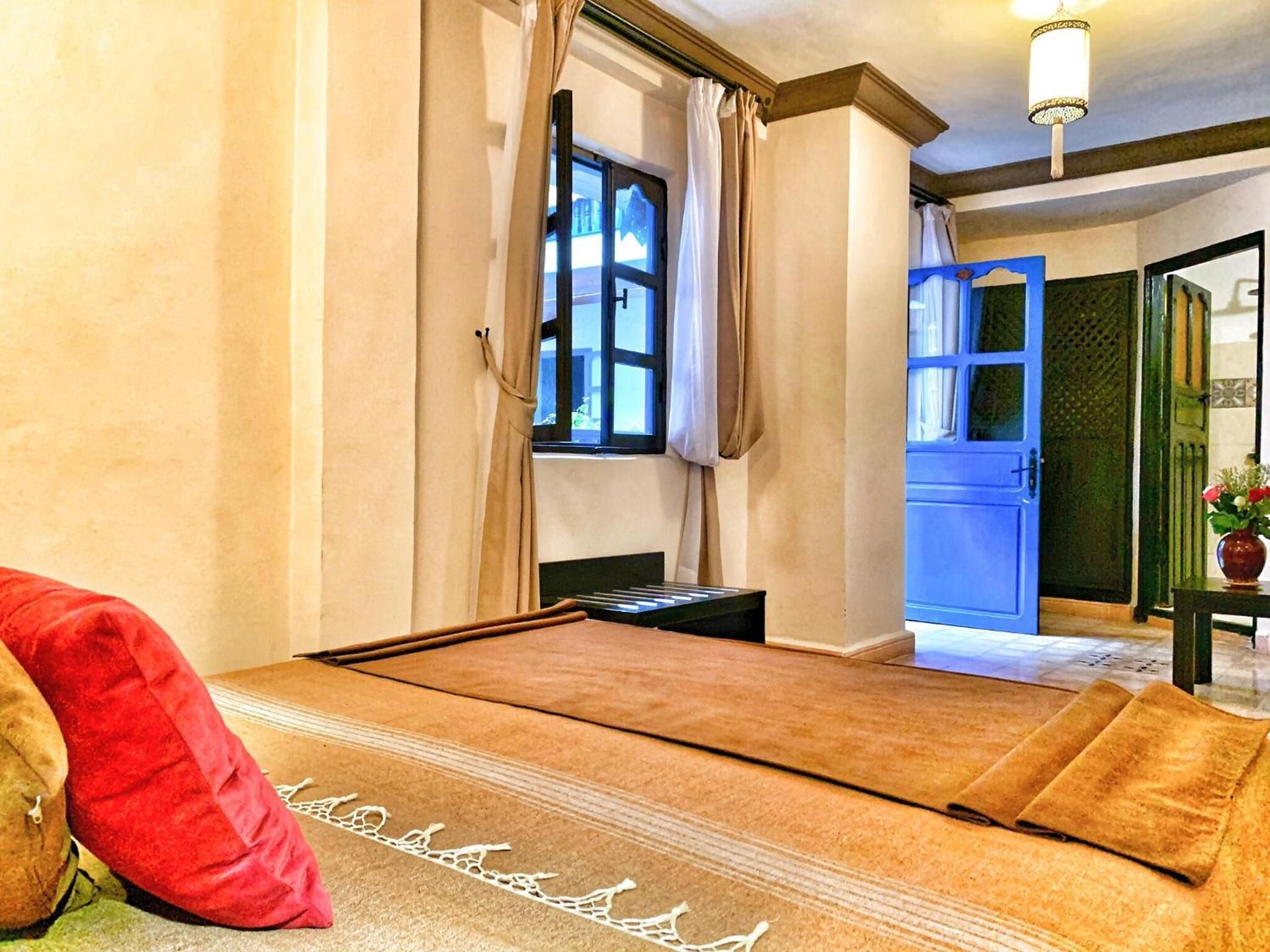 A room with a bed, a window with curtains, a blue door, and a small table with a vase.
