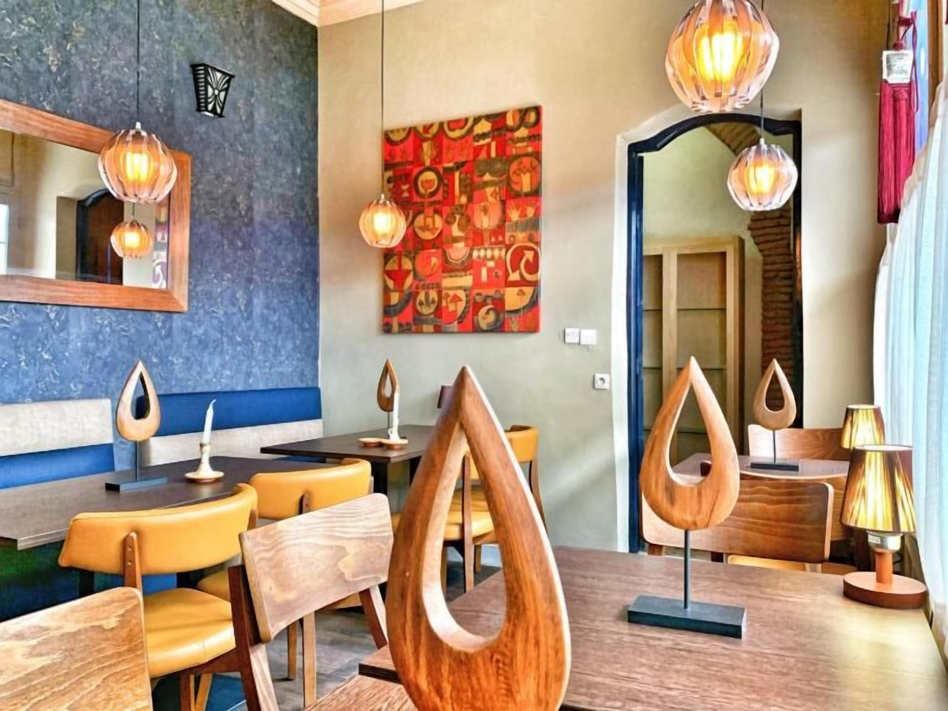 A modern dining area with wooden chairs and tables, pendant lighting, and a colorful abstract painting on the wall.
